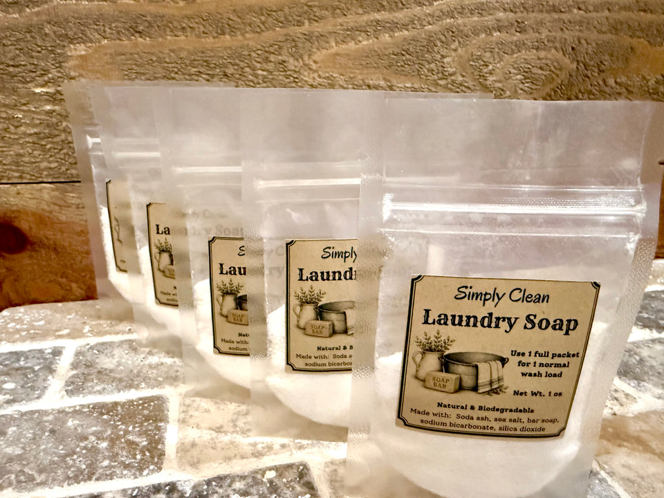 Four clear stand-up pouches of Individually packaged Simply Clean Laundry Soap arranged in a row on a stone surface against a wooden background.  Each packet is labeled as 1oz meant to be added to 1 wash load