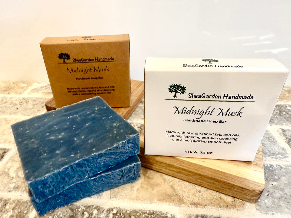 Four dark blue colored soap bars two packaged in labeled boxes with the text: Midnight Musk.  Two bars are without packaging and laying on a stone tile.

